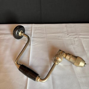 Puede incluir: Un taladro manual vintage con un mango de madera negro y un marco de metal plateado. El taladro tiene un portabrocas para sujetar brocas y un mango giratorio para su funcionamiento. La herramienta está diseñada para taladrar manualmente.