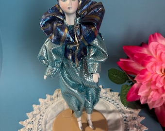 May include: A porcelain doll in a blue and gold harlequin costume, with a matching hat and a white feather. The doll stands on a beige base on a white lace doily. A pink flower is in the background.