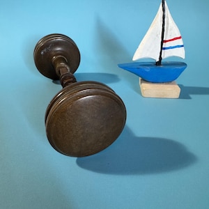 Puede incluir: Un candelabro de madera marrón oscuro y un pequeño velero azul con una vela blanca y rayas rojas y azules. El velero está montado sobre un pequeño bloque de madera. El fondo es azul claro liso.