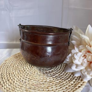 May include: A small, antique copper pot with a dark brown finish. The pot has a rounded shape with a slightly flared rim and a handle on each side. It sits on a woven, circular placemat, with a white flower in the background.