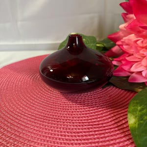 Peut inclure: Un petit vase rond en verre rouge foncé avec un col étroit. Le vase est posé sur un dessous de plat rose tissé, avec des fleurs roses et rouges et des feuilles vertes en arrière-plan. Le vase a une finition brillante.