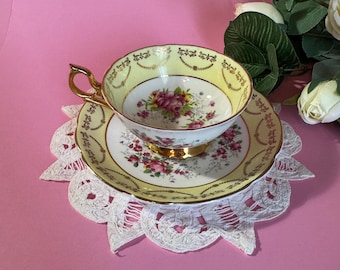 Puede incluir: Un juego de taza y platillo vintage con un diseño floral. La taza y el platillo tienen un borde amarillo con ribete dorado. La taza presenta un estampado floral en el centro. El juego se coloca sobre un tapete de encaje blanco.