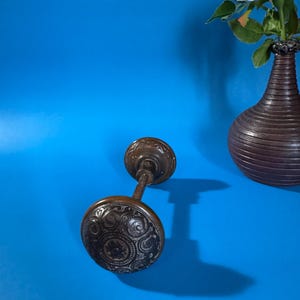 Peut inclure: Bouton de porte antique en bronze avec des motifs tourbillonnants ornés. Le bouton est relié par une fine tige à un bouton plus petit et assorti. Un vase marron avec une rose blanche est en arrière-plan sur fond bleu.