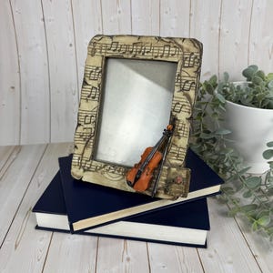 May include: A beige photo frame decorated with musical notes and a small violin. The frame sits on two dark blue books. The frame is rectangular in shape.