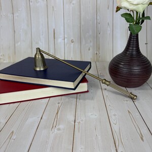 May include: A brass candle snuffer with a long handle rests on a stack of books. A dark brown vase holds a white rose. The background is a light wood grain.
