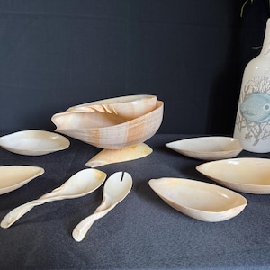 Könnte beinhalten: Eine Sammlung von beigefarbenen Servierschalen und -utensilien. Das Set umfasst ovale Schalen, Löffel und eine Serviergabel in verschiedenen Größen. Eine dekorative Flasche mit Fischmuster und Grünzeug ist ebenfalls vorhanden.