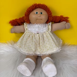 May include: A vintage doll with a round face, green eyes, and orange yarn hair styled in pigtails. The doll wears a yellow dress with a floral pattern and white lace trim, along with white socks and shoes. The doll is a collectible item.