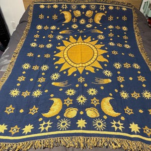Könnte beinhalten: Eine marineblaue Decke mit einem Himmelsdesign. Die Decke zeigt eine große gelbe Sonne in der Mitte, umgeben von Monden, Sternen und anderen Himmelselementen. Die Ränder sind mit gelben Quasten versehen.