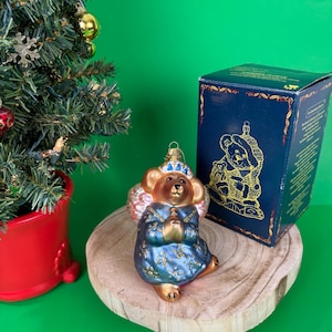 Pode incluir: Um enfeite de árvore de Natal em forma de urso, vestido com um vestido azul com detalhes dourados. O urso está ajoelhado com as patas juntas. Uma caixa azul com uma ilustração de urso dourado está atrás do enfeite. O fundo é verde.