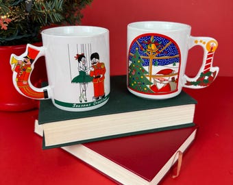 Puede incluir: Dos tazas navideñas de cerámica blanca con asas festivas. Una taza presenta un diseño de espectáculo de marionetas, la otra una escena de Papá Noel. Ambas tazas están sobre un libro verde y rojo, con un árbol de Navidad al fondo.