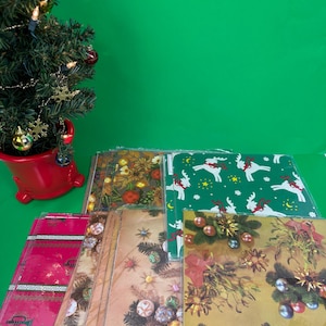 Puede incluir: Una colección de hojas de papel de regalo con temática navideña con varios diseños festivos, incluyendo renos, adornos y patrones florales. Un pequeño árbol de Navidad con adornos está en el fondo. El papel de regalo es de varios colores.