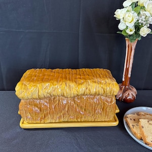 May include: A ceramic bread bin in the shape of a loaf, painted in shades of yellow and brown, sits on a yellow tray. A vase with white flowers and a plate with bread slices are also visible.