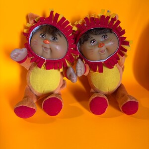 May include: Two plush dolls dressed in lion costumes. The dolls have brown hair, rosy cheeks, and orange noses. They wear red and yellow lion manes and orange and yellow outfits with yellow bellies and red feet.