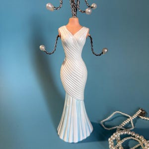 Puede incluir: Un soporte para joyas en forma de vestido blanco con cuello en V y falda acampanada, acentuado con rayas azul claro. El soporte tiene brazos y cabeza de alambre con detalles de perlas. Un collar de perlas está al lado del soporte.