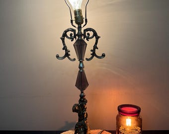 Könnte beinhalten: Eine verzierte Tischlampe im antiken Stil mit dekorativem Sockel und dunkelbraunem Schirm. Die Lampe hat eine Vintage-Ästhetik mit einer sichtbaren Glühbirne. Eine brennende Kerze in einem Glas steht neben der Lampe.