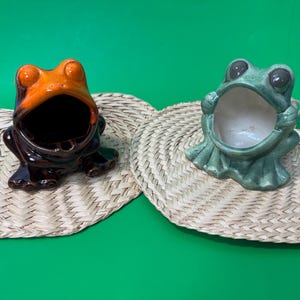 Op de afbeelding: Twee keramische asbakken in de vorm van een kikker. Eén is oranje en bruin, de andere is groen. Beide hebben open monden en zitten op geweven matten. De achtergrond is effen groen.
