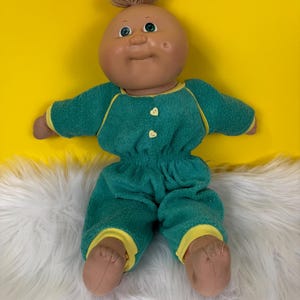 May include: A vintage doll with a tan complexion, green eyes, and a tuft of light brown hair. The doll wears a green terrycloth jumpsuit with yellow trim and two heart-shaped buttons. The doll is lying on a white fluffy surface against a yellow background.