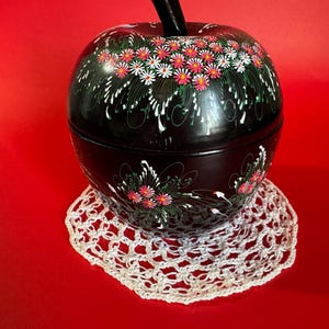 Könnte beinhalten: Ein schwarzer, apfelförmiger Behälter mit schwarzem Stiel, verziert mit einem Blumenmuster in Rosa, Weiß und Grün. Der Behälter steht auf einer weißen, gehäkelten Deckchen vor rotem Hintergrund. Der Behälter ist wahrscheinlich aus Metall oder ähnlichem Material.