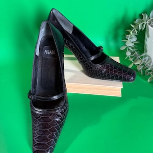 Peut inclure: Escarpins noirs en cuir avec bout carré et texture imitation reptile. Les chaussures ont une bride sur le dessus et une petite fermeture à boucle. La marque "PEGABO" est visible à l'intérieur d'une chaussure. Les talons mesurent environ 5 cm.