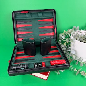 Puede incluir: Juego de backgammon negro abierto con piezas rojas y negras, dados y dos cubiletes negros. El interior es verde y rojo, con cierre de cremallera. Un juego de mesa clásico.
