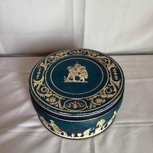 Könnte beinhalten: Runde, teal-farbene Dose mit Deckel, verziert mit goldenen Reliefmotiven und Ornamenten. Die Seiten der Dose weisen ebenfalls goldfarbene Figuren auf. Diese Vintage-Dose eignet sich zur Aufbewahrung und hat ein dekoratives Design.
