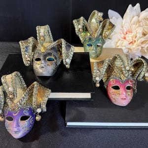 Pode incluir: Quatro máscaras venezianas decorativas com elaborados adereços de cabeça em ouro e tecido estampado. As máscaras têm rostos de cores diferentes, incluindo roxo, rosa, verde e prata, e são exibidas em superfícies pretas.