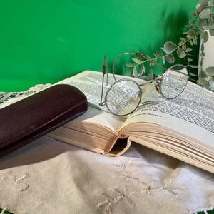 Peut inclure: Un livre ouvert avec du texte, des lunettes rondes à monture métallique et un étui à lunettes en cuir marron foncé. Les lunettes reposent sur les pages du livre, et l'étui est positionné à gauche. Le fond est vert.