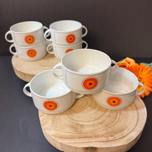 May include: A collection of white ceramic soup bowls with a retro design. Each bowl has a large orange and yellow circular graphic and a grey border. The bowls have two handles and are stacked on wooden rounds.