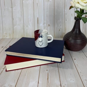 Puede incluir: Una taza blanca caprichosa con una cara de dibujos animados, ojos azules y una boca sonriente, se encuentra sobre una pila de libros. La taza tiene un diseño rojo y blanco. Un jarrón con una rosa blanca está en el fondo. Los libros son rojos y azul marino.