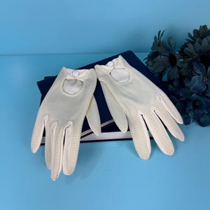 Pode incluir: Um par de luvas branco-sujo com um design perfurado. As luvas têm um fecho de botão no pulso e estão sobre uma superfície azul escura. As luvas são provavelmente de estilo vintage ou retro.