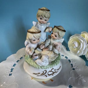 Puede incluir: Una figura de cerámica con tres ángeles con halos dorados, rodeando a un bebé en un pesebre. La base blanca tiene un borde dorado y un diseño decorativo. La figura está colocada sobre un mantel blanco.