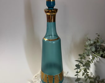May include: A teal glass decanter with a matching stopper and gold accents. The bottle has a slender shape with a wide base and a decorative gold band. The stopper is long and cylindrical. The decanter sits on a white lace doily.