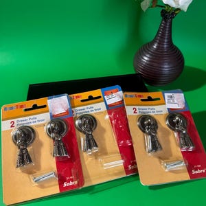 May include: Three packs of Home Time drawer pulls with a decorative design. Each pack contains two pulls and mounting hardware. The pulls are a dark metallic colour and are displayed on a green background.