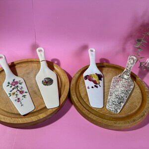 Peut inclure: Quatre pelles à gâteau décoratives avec des lames en céramique blanche et des manches en bois. Deux pelles reposent sur des plateaux de service en bois. Les lames présentent des motifs floraux, de fruits et abstraits. Le fond est rose vif.