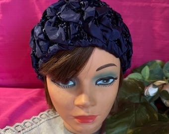 May include: A navy blue vintage hat with a textured, ruffled design. The hat is displayed on a mannequin head with brown hair, blue eye shadow, and red lipstick. The background is a bright pink fabric.