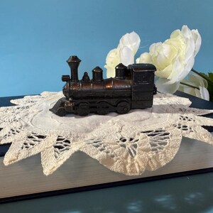 May include: A miniature antique-style train engine, in shades of brown, sits atop a cream-colored lace doily. The train is placed on a navy blue book, with white flowers in the background.