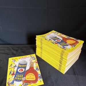 May include: A stack of yellow Bee Hive Golden Corn Syrup advertising flyers. Each flyer features a vintage illustration of a syrup bottle and can, with text in both English and French. The flyers are on a black surface.