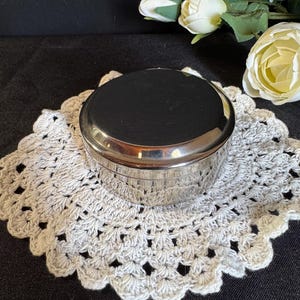 May include: A small, round, silver-colored container with a lid, resting on a white, crocheted doily. The container has a shiny, reflective surface. White roses and buds are in the background.