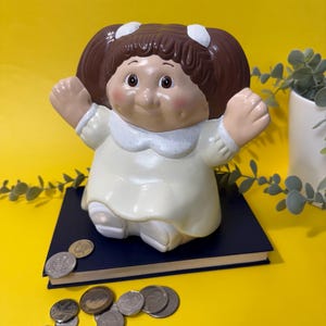 May include: A vintage ceramic doll-shaped piggy bank with brown hair and white bows. The doll wears a cream-coloured dress and sits on a navy blue book. Coins are scattered on a bright yellow surface.