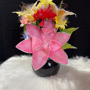 Può includere: Una vivace composizione floreale con un grande fiore rosa in primo piano, circondato da fiori gialli, rossi e bianchi. I fiori sono in un vaso nero, su sfondo nero.