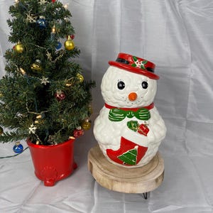 Puede incluir: Un tarro de galletas de cerámica con forma de muñeco de nieve con sombrero rojo, pajarita verde y diseño de calcetín. Un pequeño árbol de Navidad con adornos está en una maceta roja.