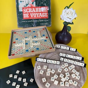 Pode incluir: Conjunto de jogo de tabuleiro Scrabble de Voyage vintage. A imagem mostra o tabuleiro do jogo, as peças de letras e a caixa com o título do jogo em vermelho. Uma rosa branca num vaso castanho também está na imagem.
