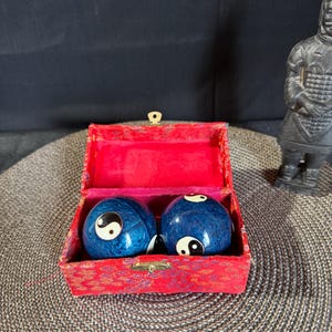 Può includere: Due sfere cinesi blu, dall'aspetto marmorizzato, con simboli yin-yang, sono in una scatola rossa foderata di tessuto. Una piccola statua scura è visibile sullo sfondo.
