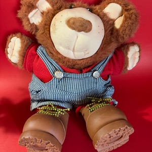 Op de afbeelding: Een bruine teddybeer met een rood shirt en een blauw-wit gestreepte overall. De beer heeft grote bruine laarzen met gele veters. Het gezicht van de beer is crèmekleurig met een bruine neus en een vriendelijke uitdrukking.