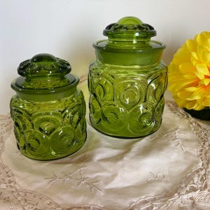 May include: Two green glass jars with lids. The jars feature a textured pattern of circles and crescent shapes. A large yellow flower is in the background. The jars are on a cream-coloured, embroidered cloth.