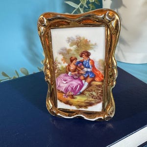 May include: A small, rectangular porcelain picture frame with a gold-coloured, ornate border. The framed image depicts a couple in period clothing, with a man in blue and a woman in pink. The artwork is signed "Fragonard".