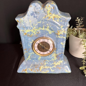 May include: A vintage ceramic clock with a marbled blue, yellow, and gold design. The clock has a round face with gold trim and Roman numerals. The clock is a decorative item.