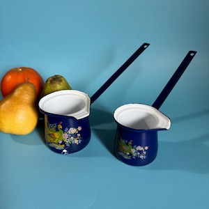 May include: Two navy blue enamel coffee pots with long handles and white interiors. Each pot features a peacock and floral design. A pear, orange, and another pear are next to the pots.