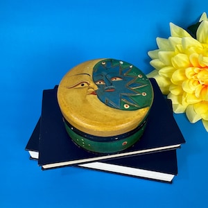 Puede incluir: Una caja redonda de madera con tapa que presenta un diseño celestial. La tapa representa una luna amarilla y un sol azul abrazándose. La caja es verde y amarilla, apoyada sobre dos libros azul oscuro, con una flor amarilla en el fondo.