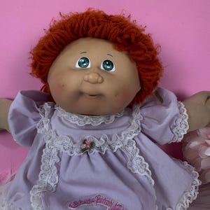 Puede incluir: Muñeca vintage Cabbage Patch Kids con cabello rizado rojo y ojos azules. La muñeca viste un vestido lavanda con ribete de encaje blanco y un pequeño detalle floral. La muñeca tiene mejillas rosadas.